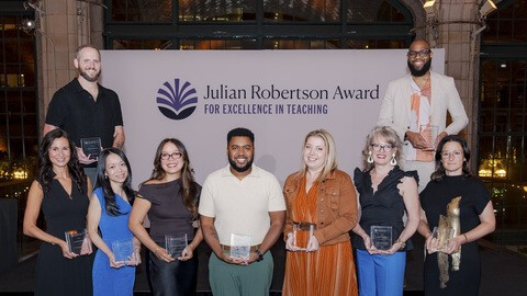 Nine people stand on stage holding glass awards in front of a banner proudly displaying the Julian Robertson Award for Excellence in Teaching.